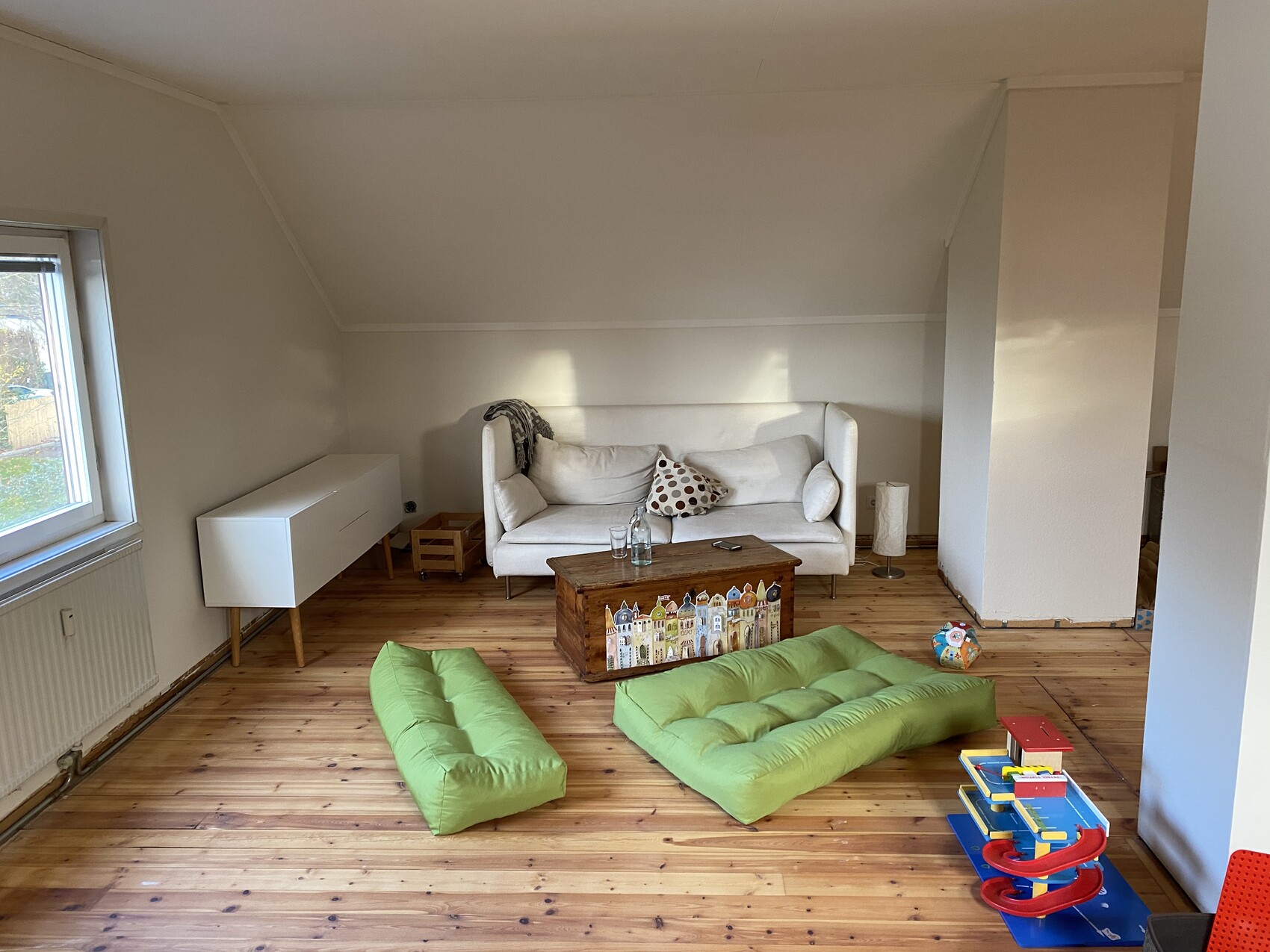 Wohnzimmer mit weißer Couch, Holzboden, zwei grüne Sitzkissen und bunte Spielzeuge.