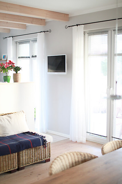 Wohnzimmer mit hellgrauen Wänden, weißen Vorhängen, Wand-TV, Korbsessel und Pflanze auf Kommode