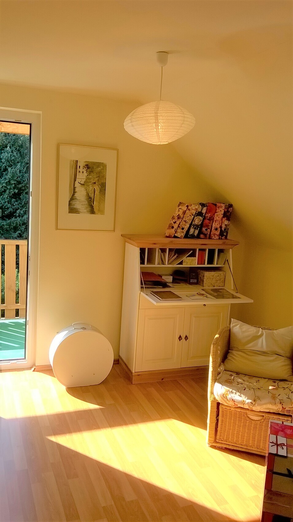 Helles Wohnzimmer-Eck mit weißem Sekretär, Korbsessel, runder Lampenschirm und Balkontür.