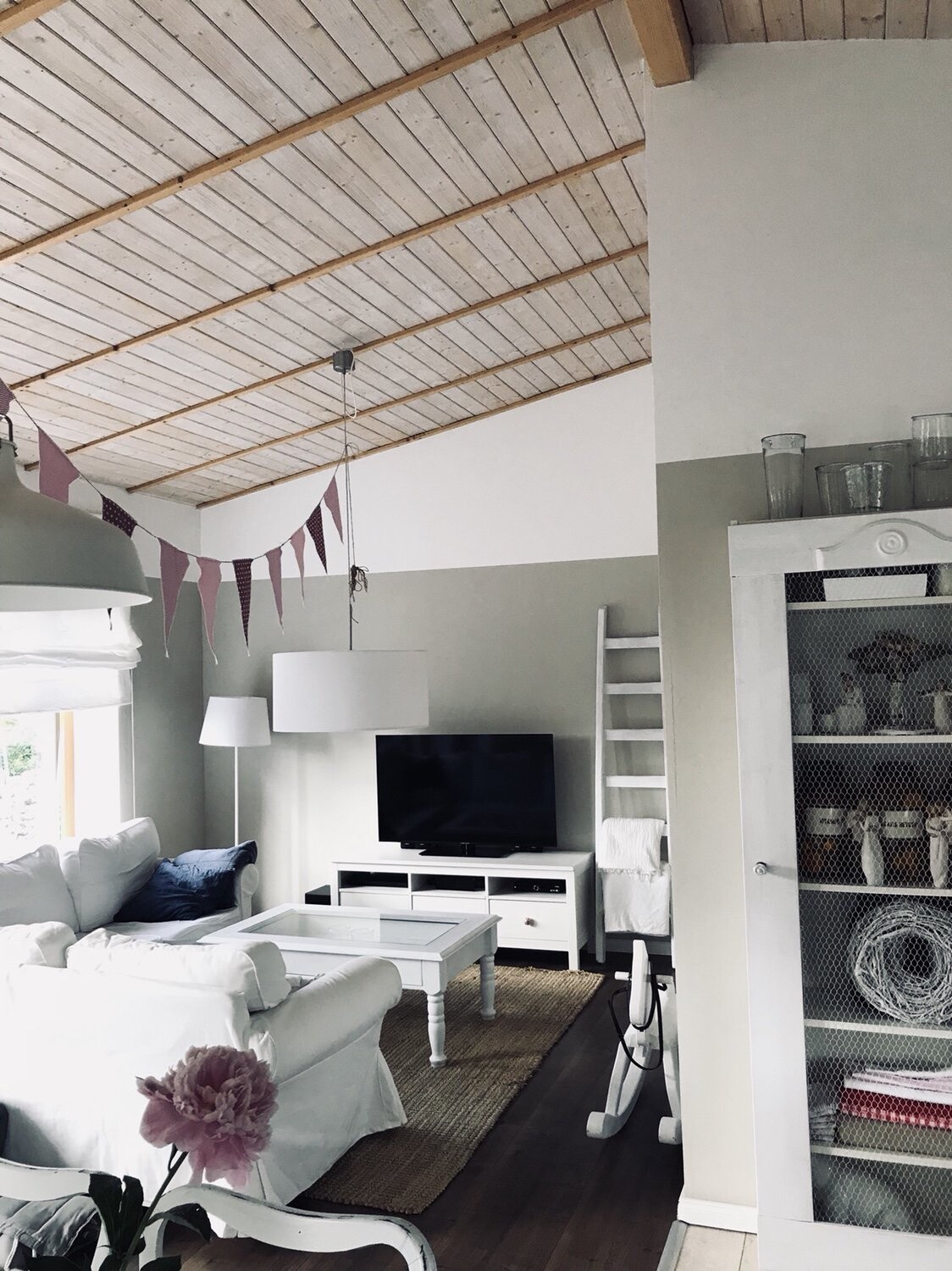 Wohnzimmer mit weißen Sofas, TV, Glasvitrine, Holzdecke, rosa Girlande und Blüte im Vordergrund