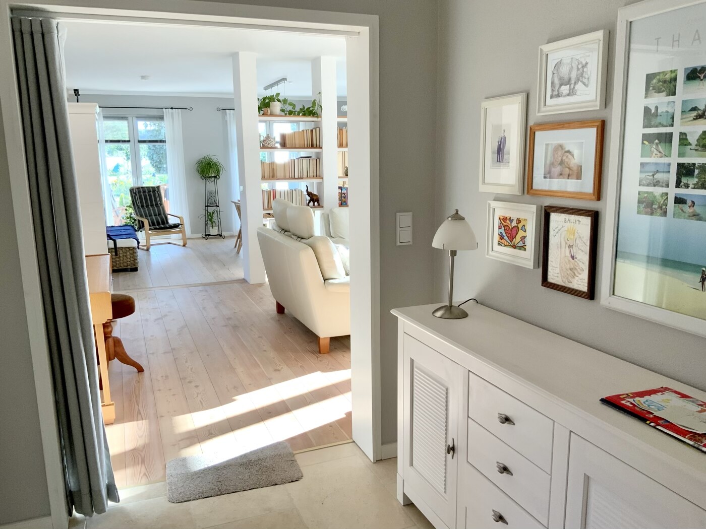 Heller Wohnraum mit weißem Sofa, Bilderrahmen-Galerie an der Wand und Bücherregal im Durchgang.