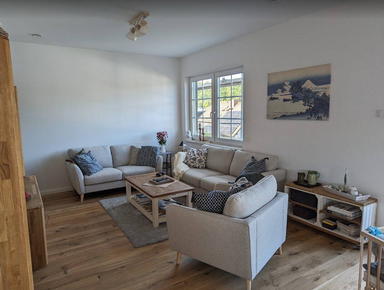 Helles Wohnzimmer mit L-förmiger Sofa-Gruppe, Holzboden, Couchtisch, Fenster mit Aussicht, Wandbild.