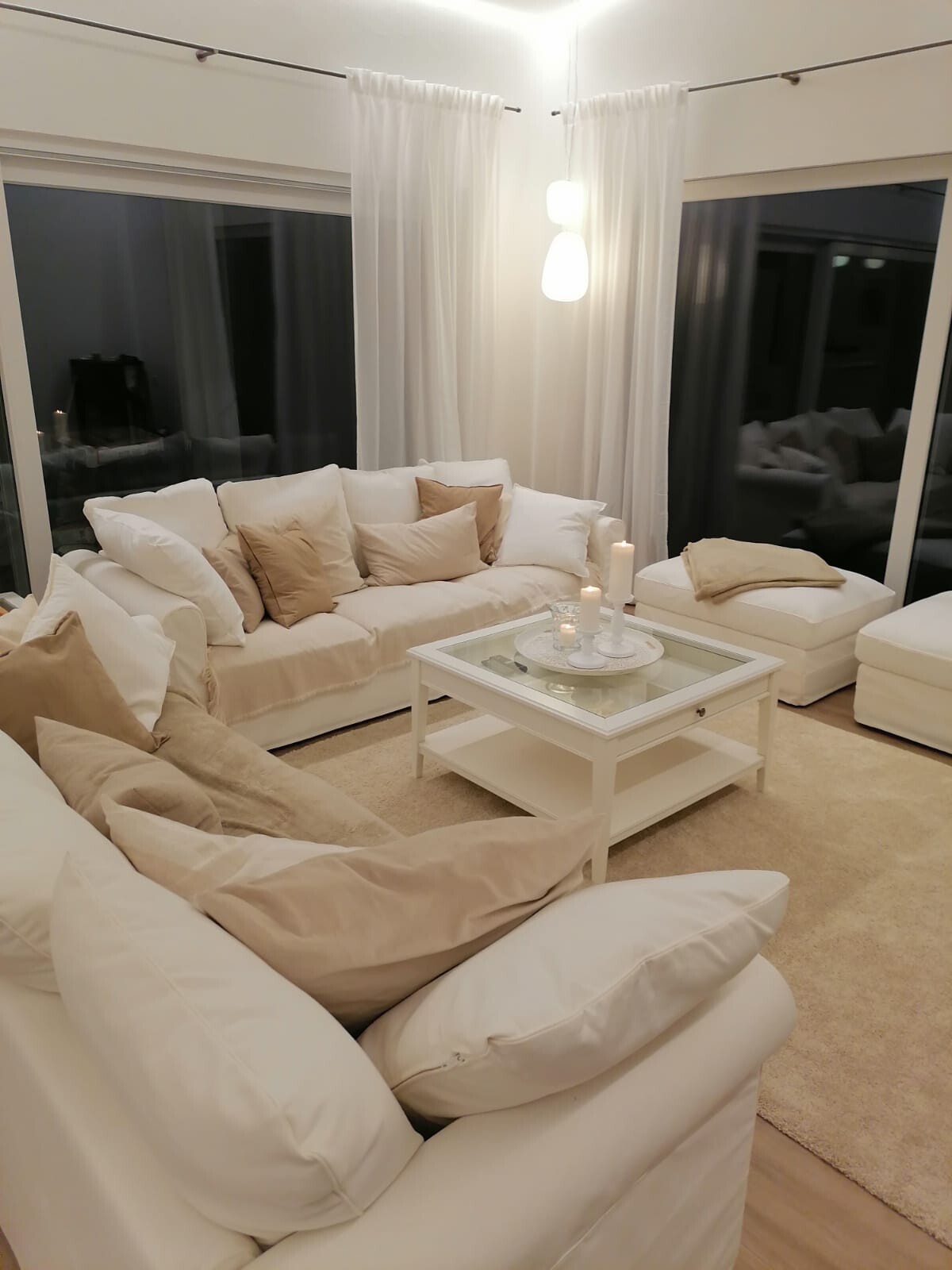 Helles Wohnzimmer mit weißen Sofas, beige Kissen, Glas-Couchtisch, Kerzenlicht und großen Fenstern.