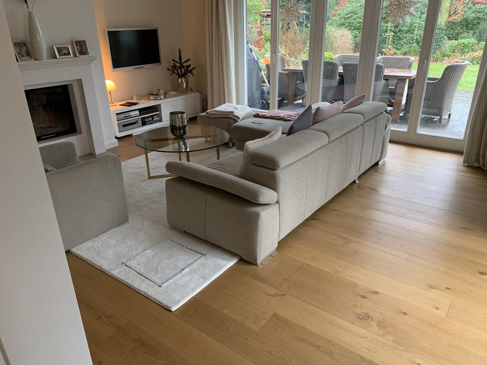 Helles Wohnzimmer mit beigem Ecksofa, Glas-Couchtisch, Kamin, TV, Blick auf Terrasse und Garten.