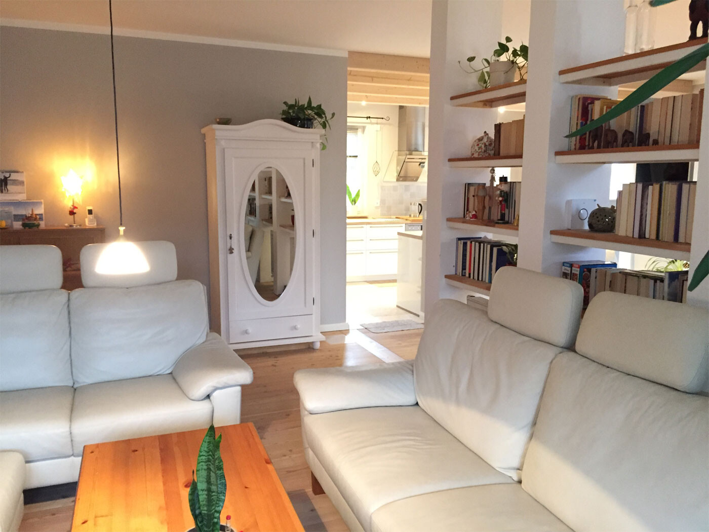 Heller Wohnzimmer mit weißen Ledersofas, weißem Schrank und Bücherregal; offene Küche im Hintergrund.