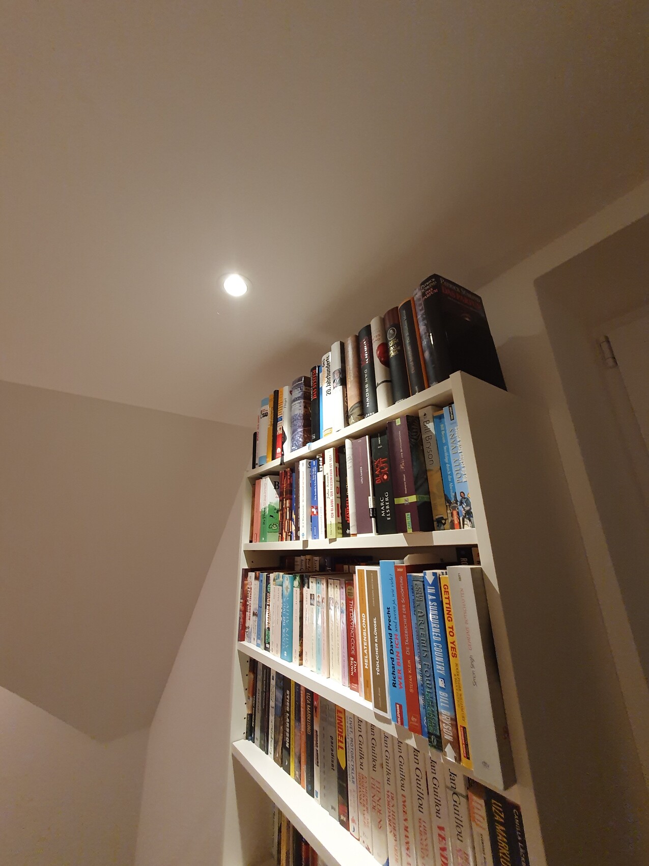 Weißes Bücherregal voller Bücher in einer Wohnzimmerecke, Deckenlicht darüber.