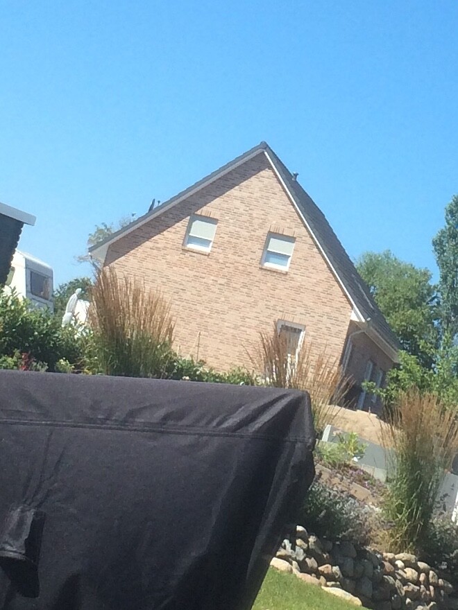 Ziegelhaus mit Satteldach; vorn eine schwarze Abdeckung über Gartenmöbel, blauer Himmel.