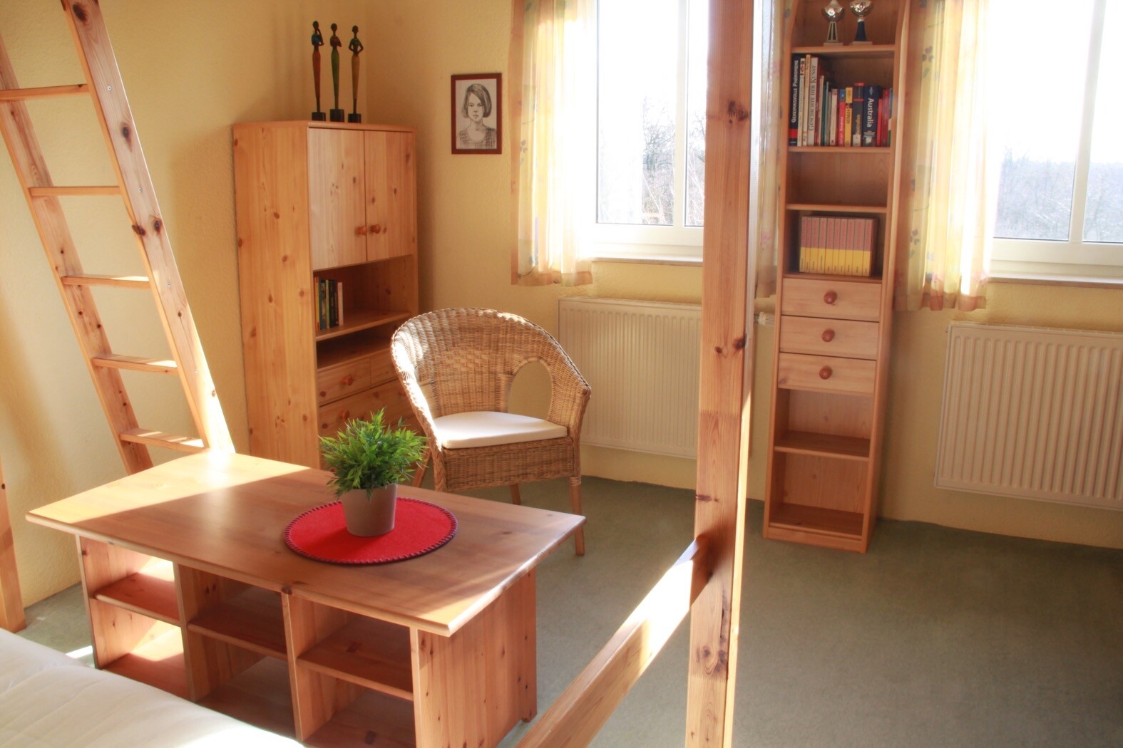 Heller Holztisch mit Pflanze, Korbsessel, Bücherregal, Leiter; Fenster mit Vorhängen.