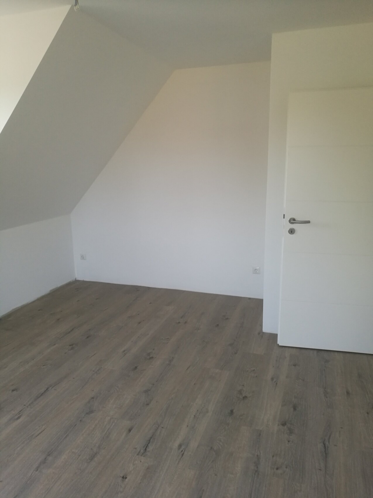 Leerer Raum mit weißen Wänden und Holzboden; links geneigtes Dach, rechts geschlossene weiße Tür.