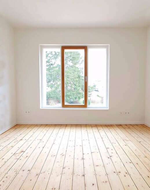 Helles leeres Zimmer mit Holzparkettboden und großem Fenster mit Baumansicht.