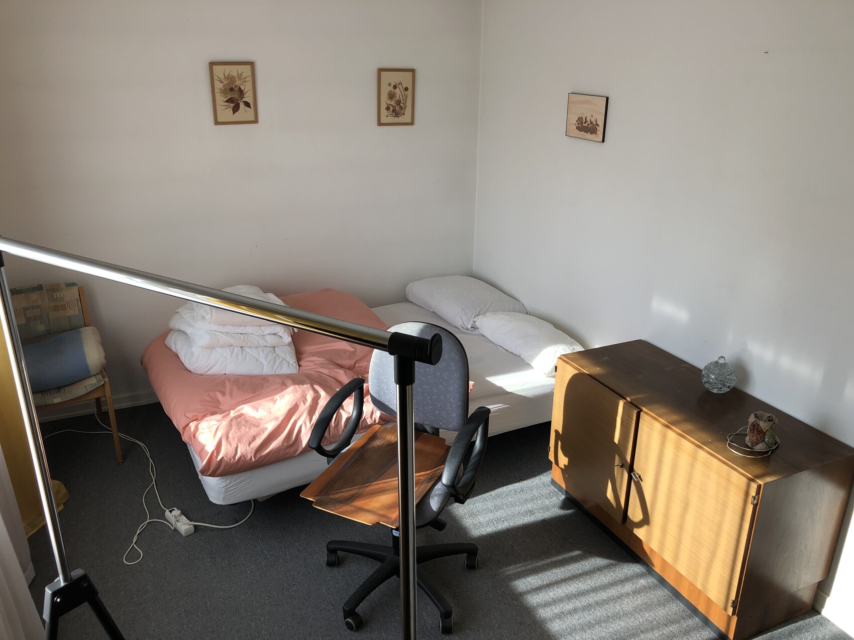 Zwei Einzelbetten in einem einfachen Zimmer; links rosa Decke, rechts weiße Bettwäsche, Holzkommode.