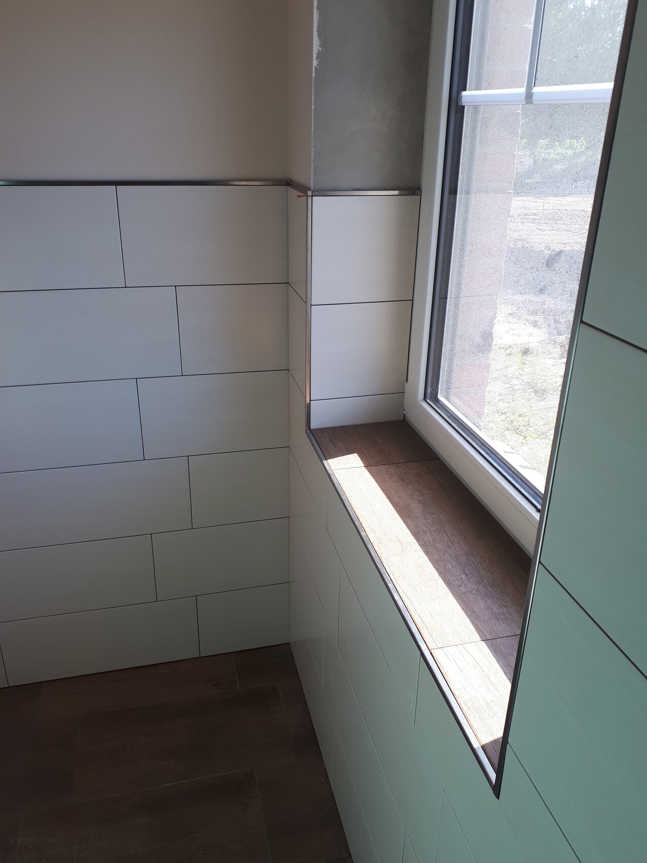 Heller Badezimmerbereich mit mintgrünen Rechteckfliesen, Fensterbank aus Holz und dunklem Boden.