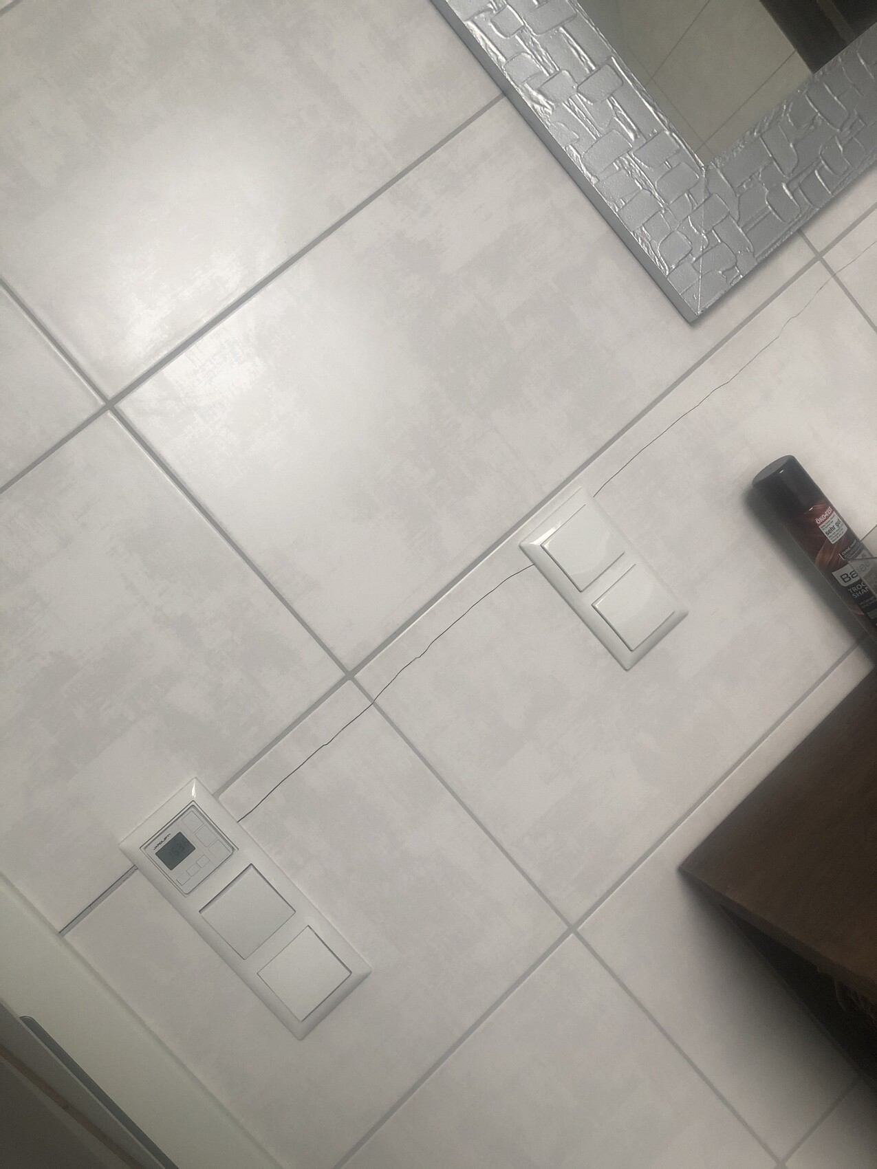 Graue Fliesenwand mit zwei Schaltern, Thermostat links, Spiegel mit Mosaikrahmen; Sprühdose rechts.
