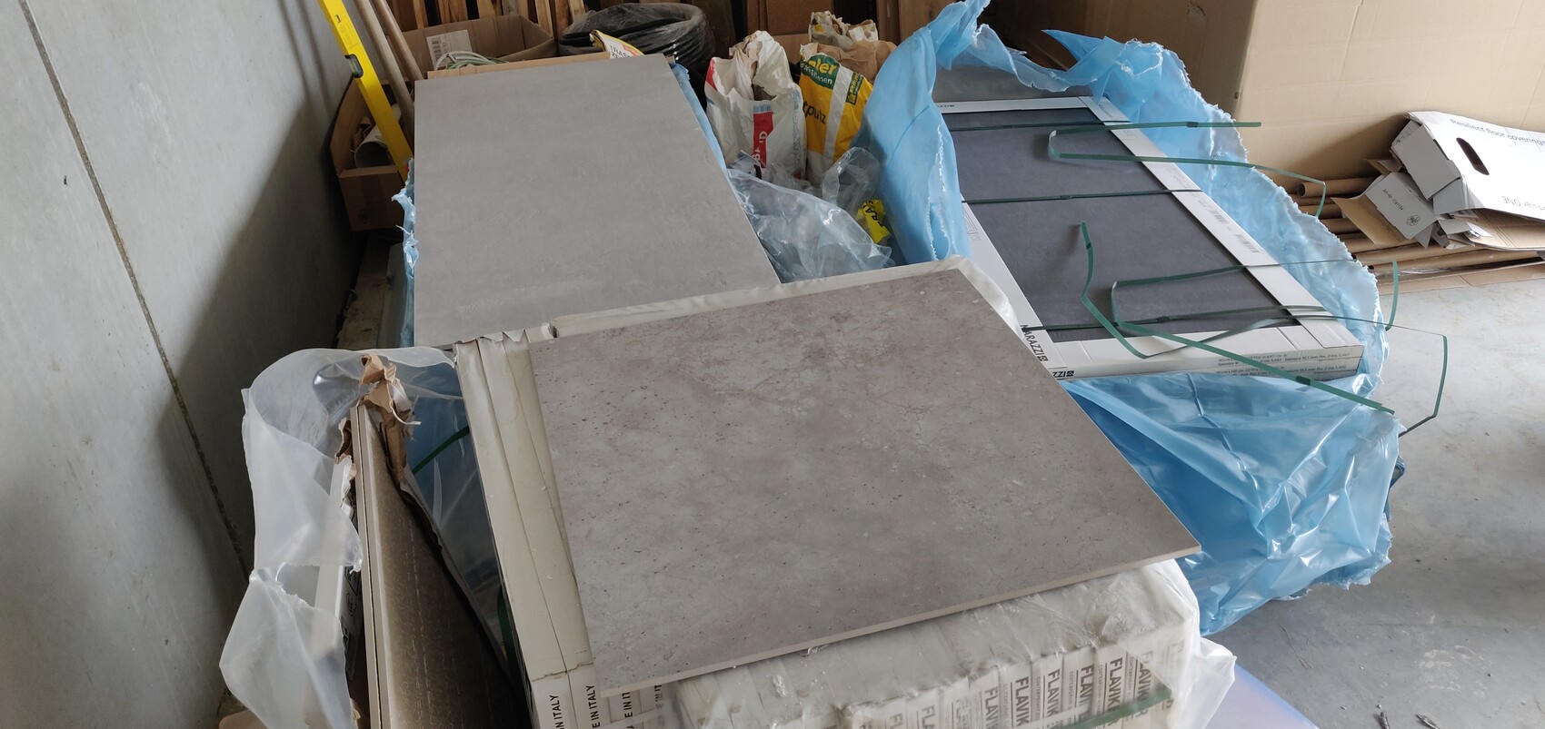 Große Fliesenplatten liegen auf Paletten, umgeben von Verpackung und blauer Folie im Lager.