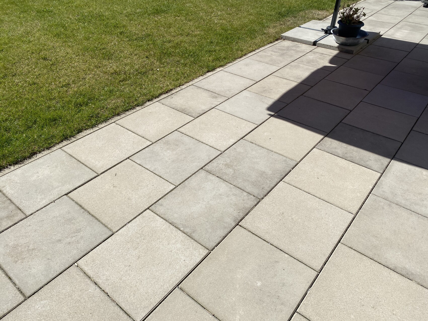 Gepflasterter Patio aus grauen Rechteckplatten, Gras links, Pflanzkübel rechts, Schatten über Steinen.