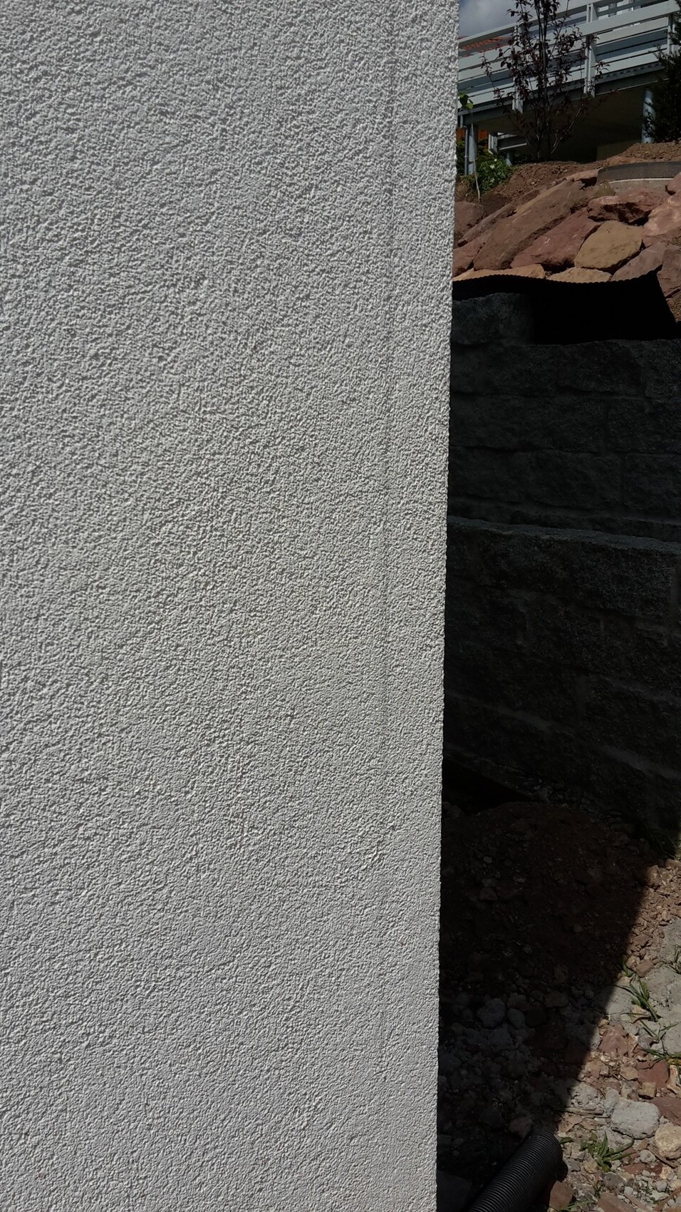 Nahaufnahme einer grob texturierten hellgrauen Außenwand; rechts dunkle Steinmauer mit Erdreich.
