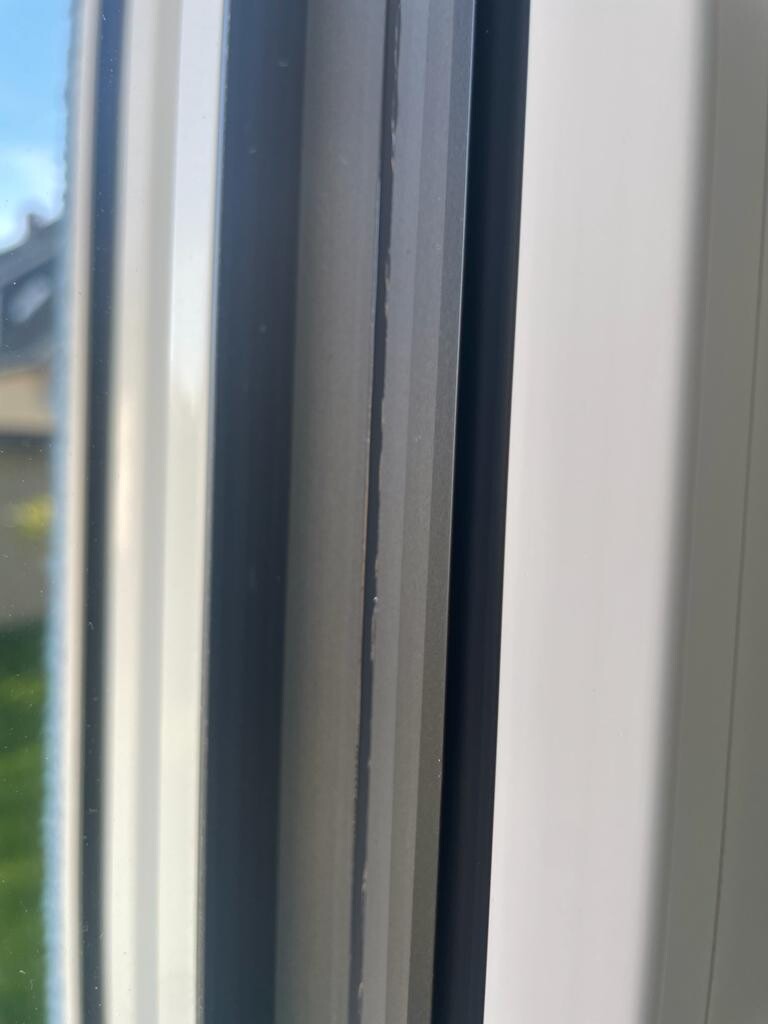 Nahaufnahme eines Fensterrahmens: dunkelgrauer Innenrahmen neben hellem weißen Rahmen, draußen unscharf