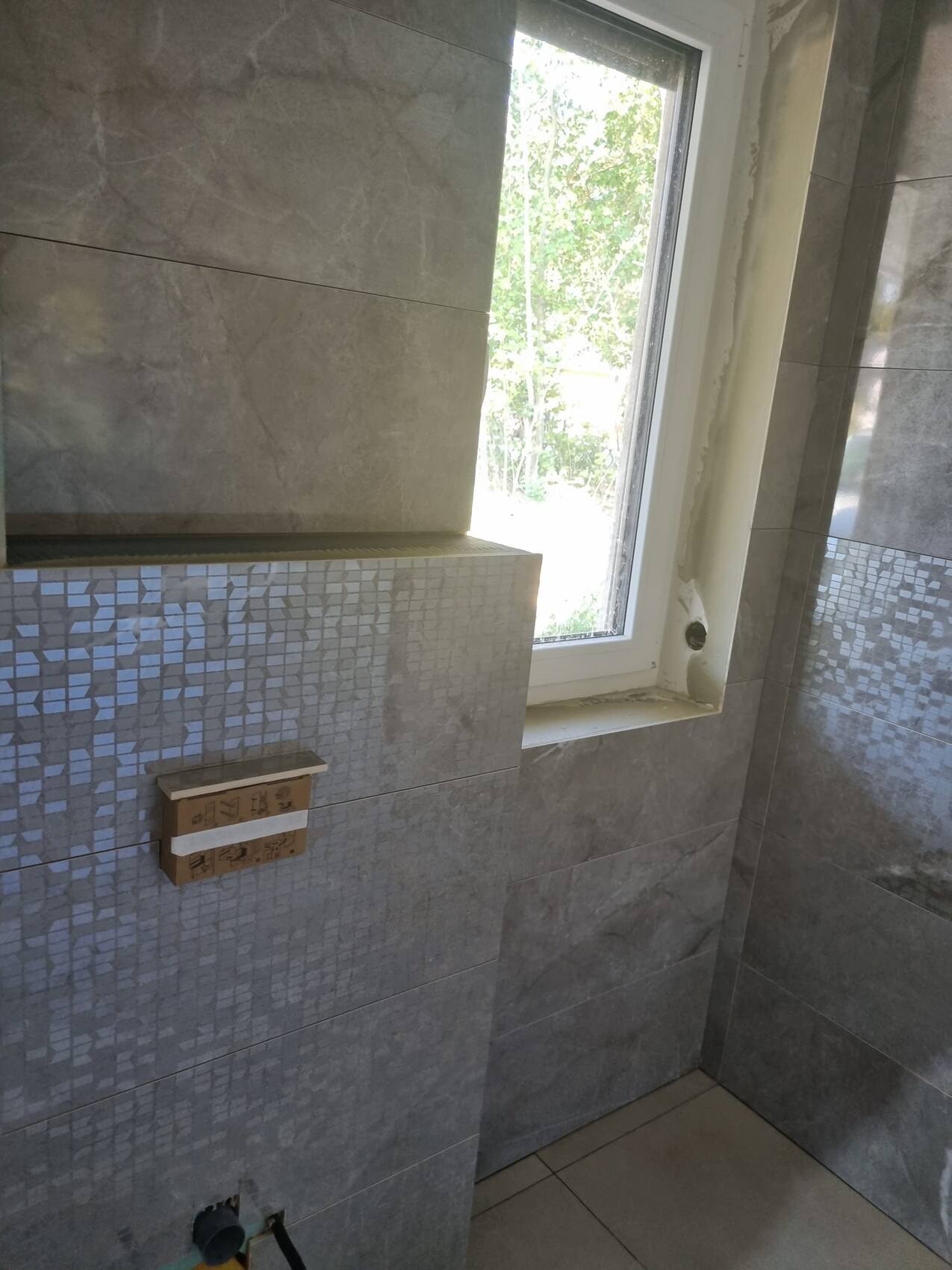 Badezimmerwand mit grauen Fliesen und Mosaikband, Fenster mit Blick ins Grün draußen.