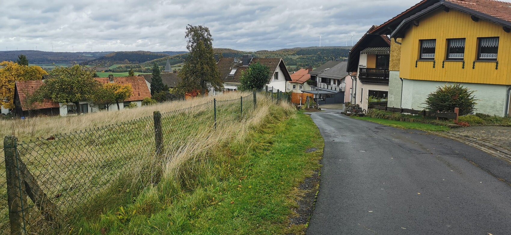 Ländliche Dorfstraße mit gelbem Haus rechts, Zaun links, Grasland und Windräder im Hintergrund.