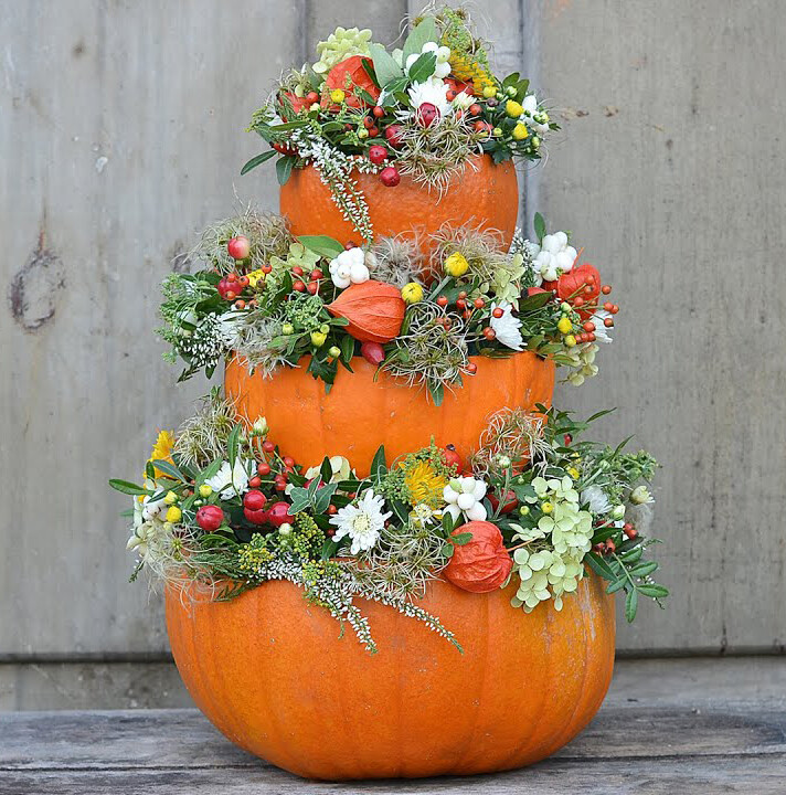 Drei orange Kürbisse stapeln sich als Herbstdekoration, verziert mit bunten Blumen und Grün.