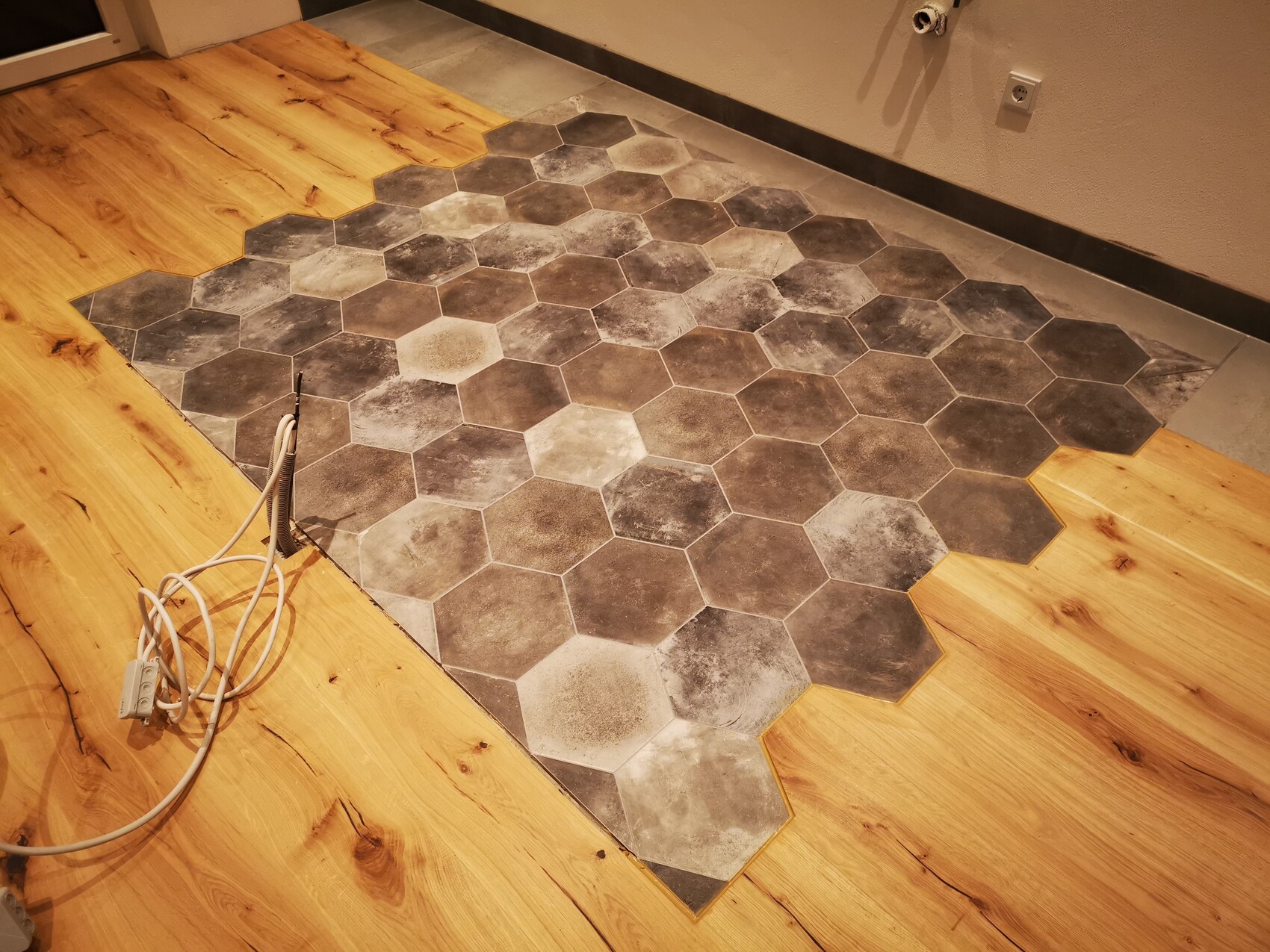 Boden mit grau-schwarzen Hexagonfliesen, umlaufender Holzboden; Kabel liegen darauf.