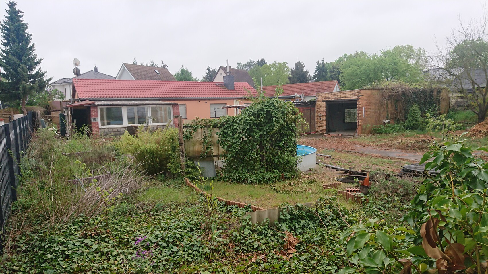 Garten mit überwucherter Vegetation, blauem runden Pool und Backsteingarage im Hintergrund.