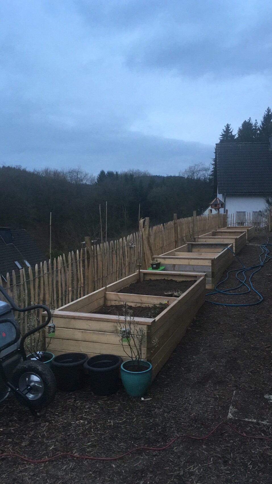 Mehrere rechteckige Hochbeete aus Holz mit Erde, Zaun und Haus im Hintergrund.