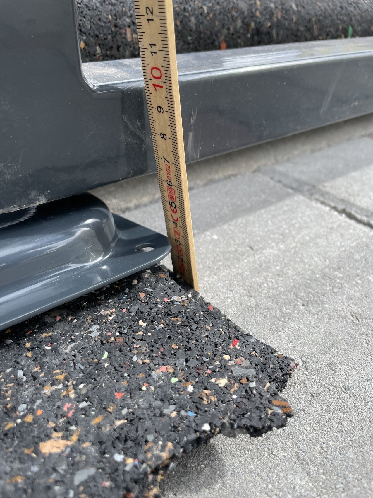 Holz-Zollmaßstab steht senkrecht neben Asphaltkante und grauer Metallkante; ca. 12 Zoll.