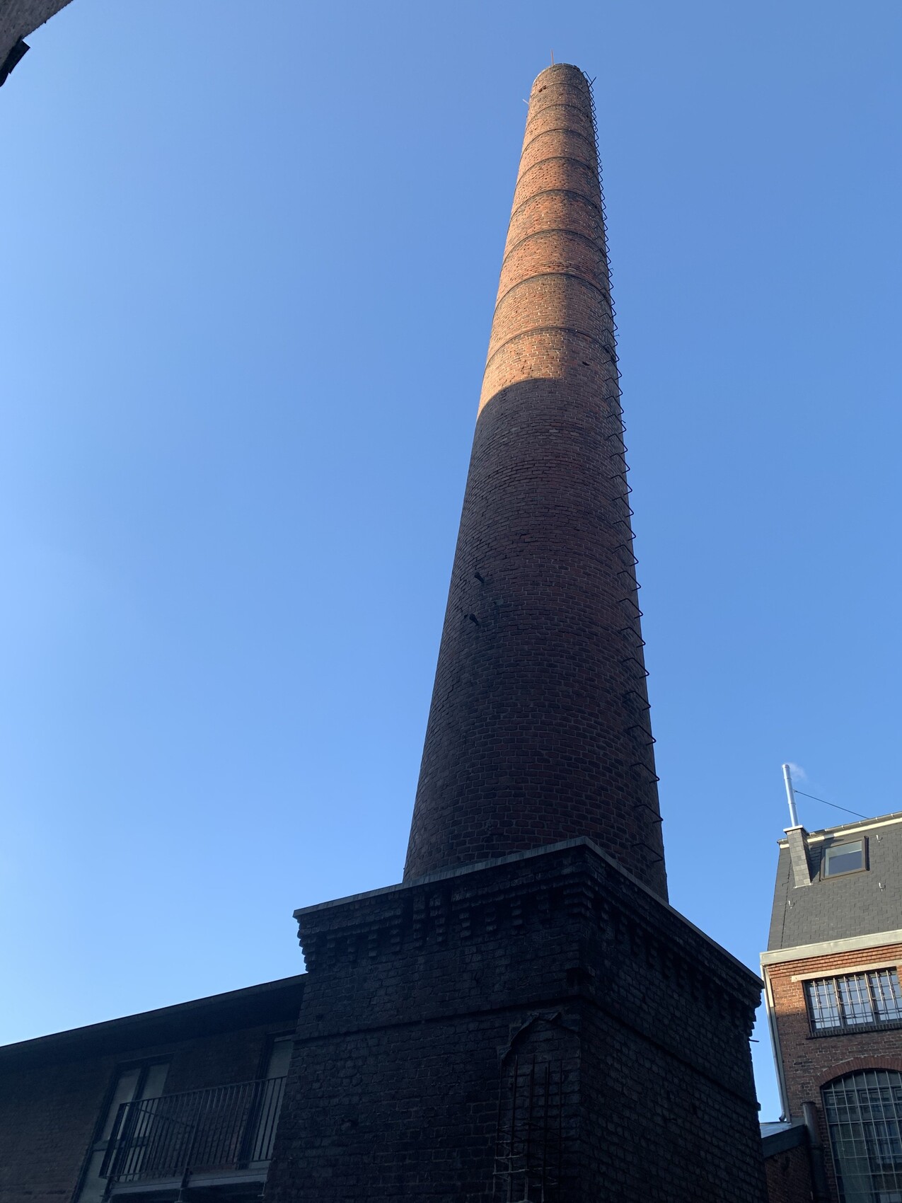 Hoher Backstein-Schornstein neben einer Fabrik, äußere Leiter am Rand, blauer Himmel.