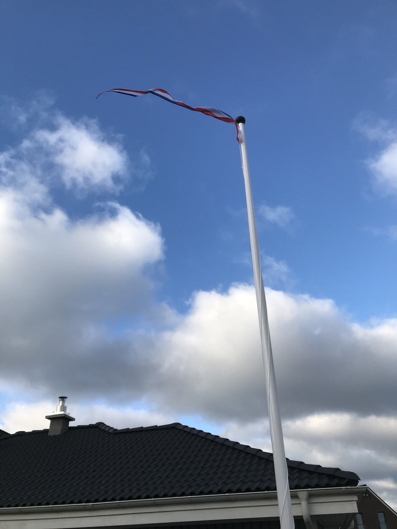 Hohe Flaggenstange mit rotem und weißem Band weht vor dem Hausdach gegen blauen Himmel.