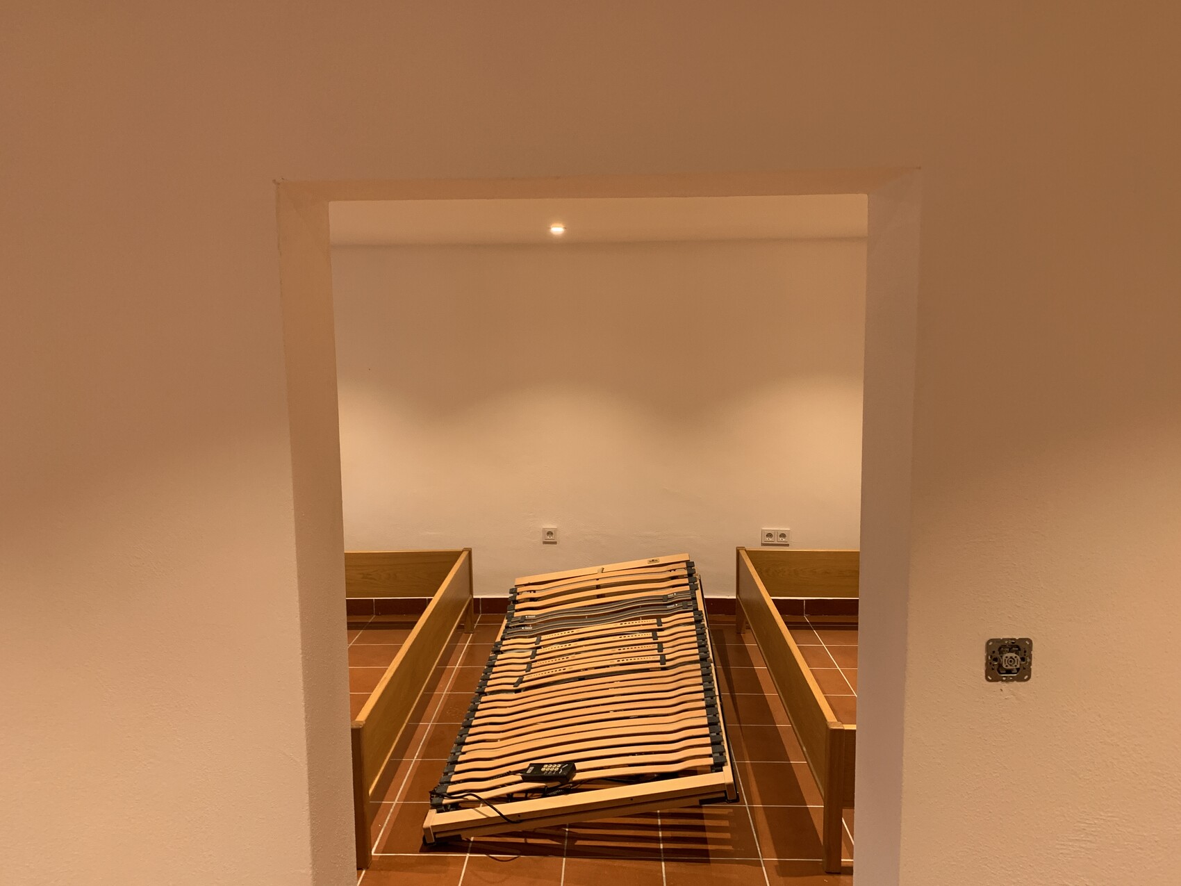 Bettgestell mit Lattenrost, seitliche Holzleisten, auf Fliesenboden, warmes Licht.