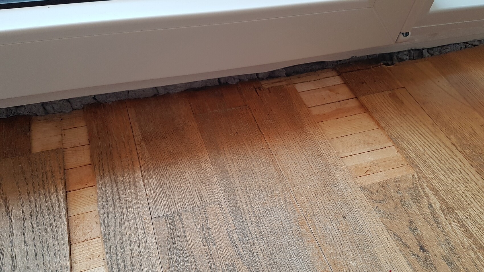 Weißer Heizkörperunterkasten über unregelmäßigem Holzboden mit grauer Dämmung.