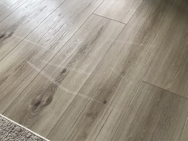 Heller Holzboden mit Planken, weißen Kreideflecken und Teppichkante unten links.