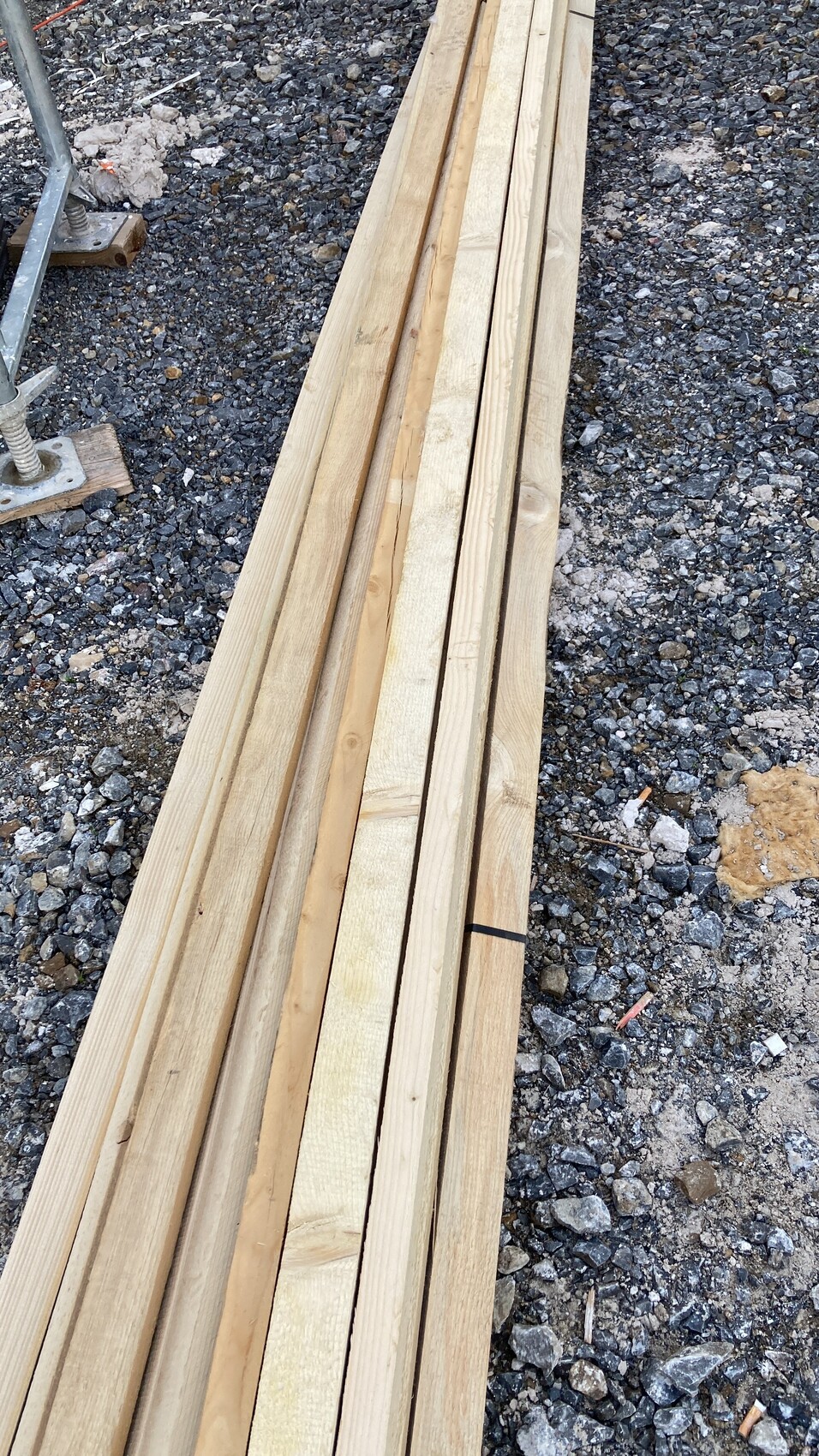 Mehrere lange Holzbretter liegen auf Kies auf einer Baustelle, rechts ein Metallgerüst.
