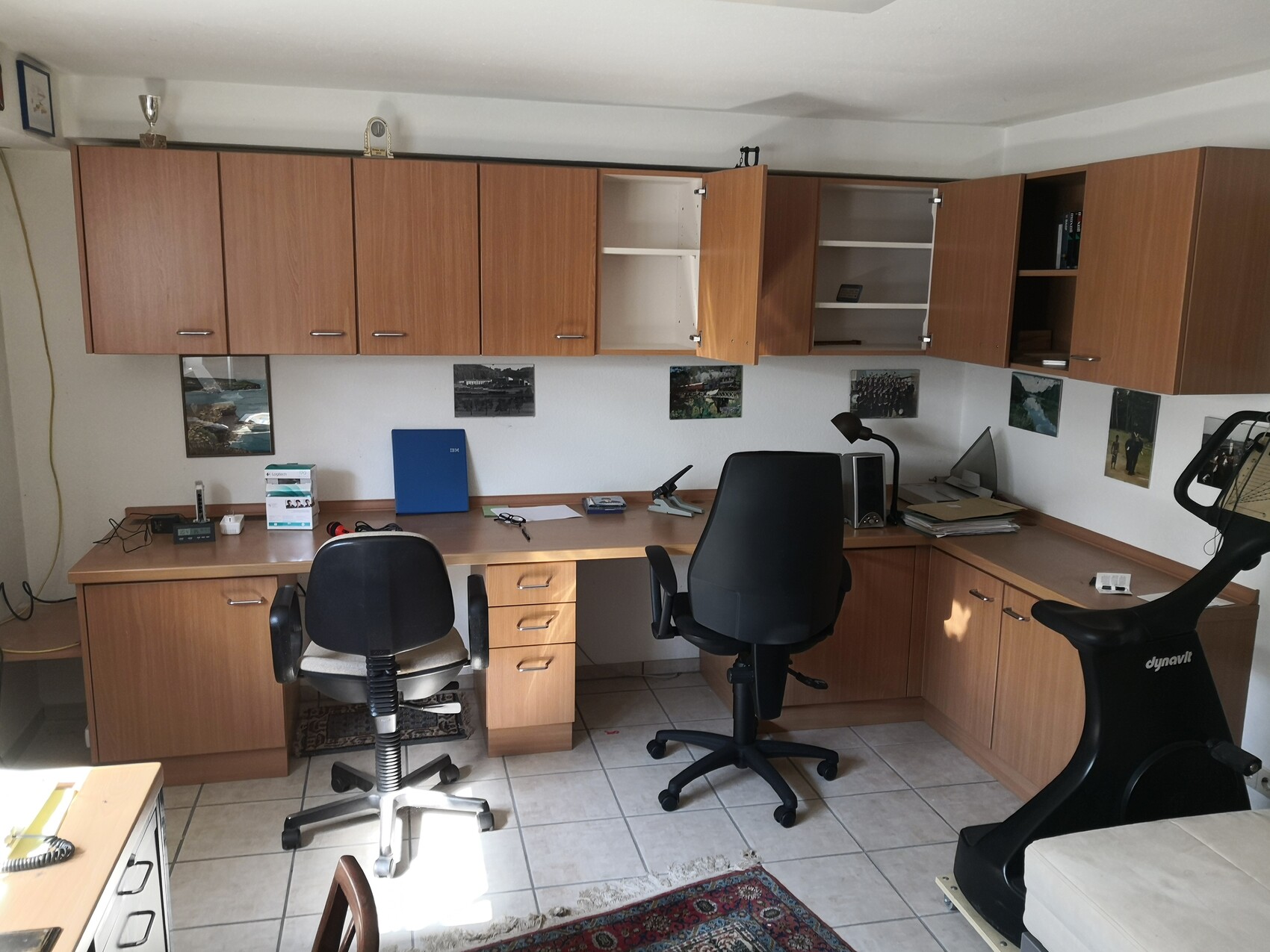Arbeitszimmer mit Holzschränken entlang der Wand, Schreibtisch, zwei Bürostühle und Fitnessgerät.