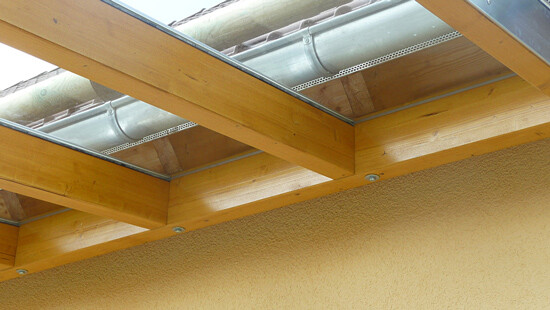Holzpergola mit Balken, Glasfenstern dazwischen über gelber Wand; obere Metallrinne sichtbar.
