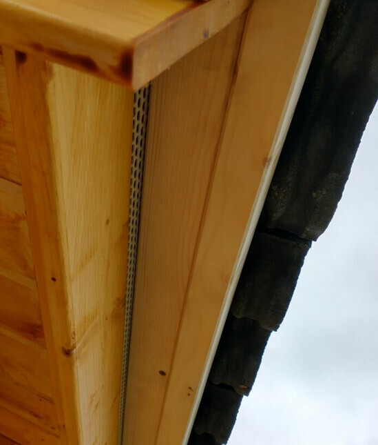 Nahaufnahme einer Holzkonstruktion am Dachrand mit Metallstreifen und dunklen Dachziegeln.