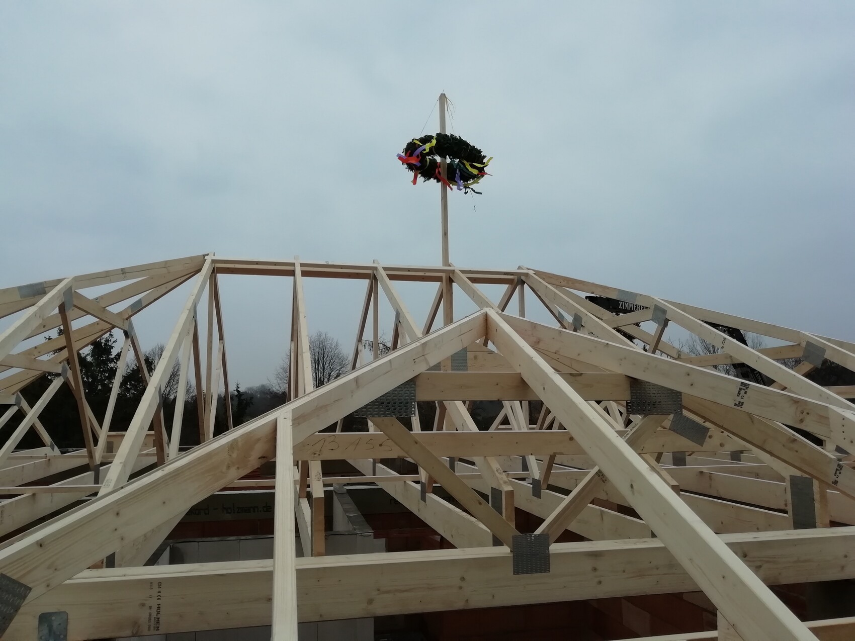 Holz-Dachstuhlbau mit zentralem Mast und bunten Bändern unter wolkigem Himmel