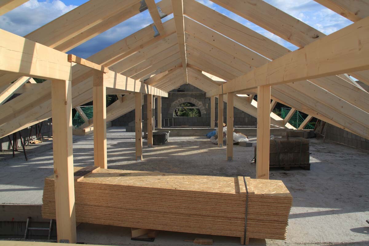 Holz-Dachstuhlrahmen auf einer Baustelle; vorne gestapelte Holzbretter, Betonboden sichtbar.