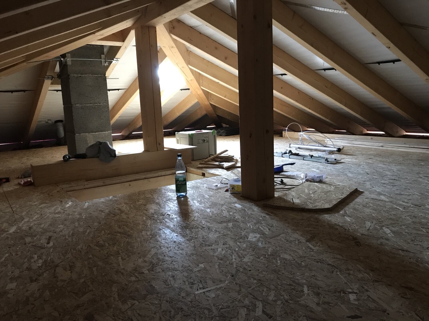 Unfertiger Dachboden mit Holzsparren, Betonsäule, Werkzeugen, Wasserflasche und OSB-Boden.