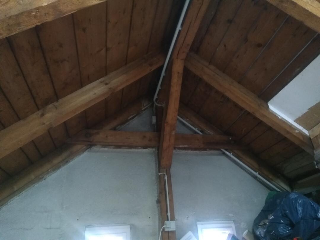 Dachboden mit Holzbalken-Dachstuhl, sichtbare Rohrleitungen und blauer Müllsack in der Ecke