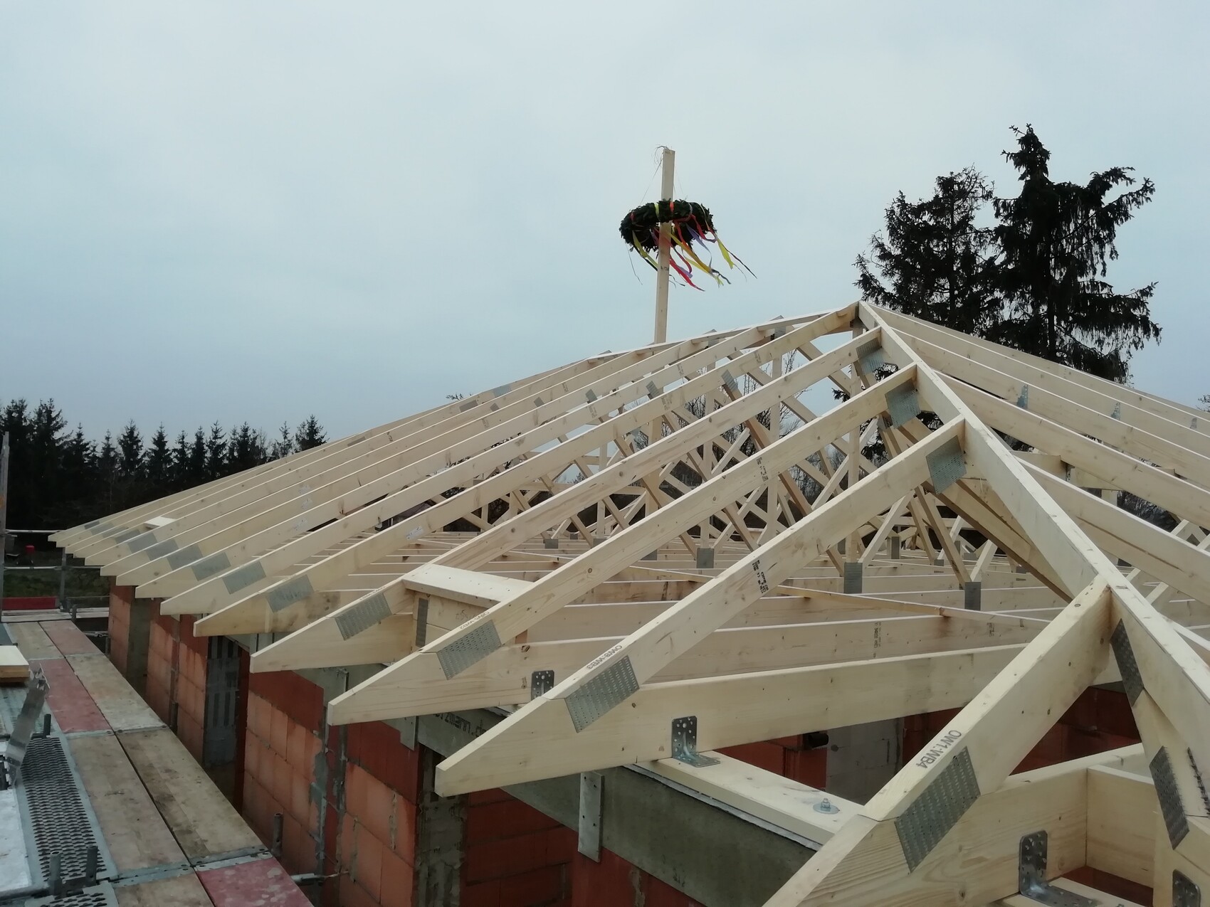 Dachstuhl aus Holz über einem Ziegelbau; Mast mit Kranz und bunten Bändern am First.