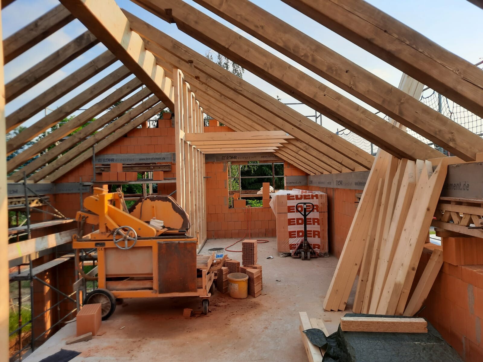 Innenansicht einer Baustelle: Dachkonstruktion aus Holzbalken, Ziegelwänden und Baumaterial.