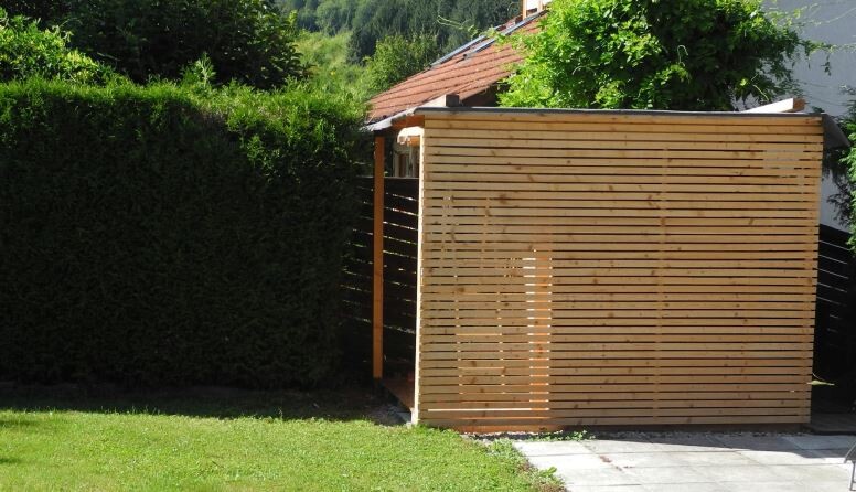 Kleiner Garten-Holzschuppen mit horizontalen Latten, neben grüner Hecke und gepflastertem Weg.