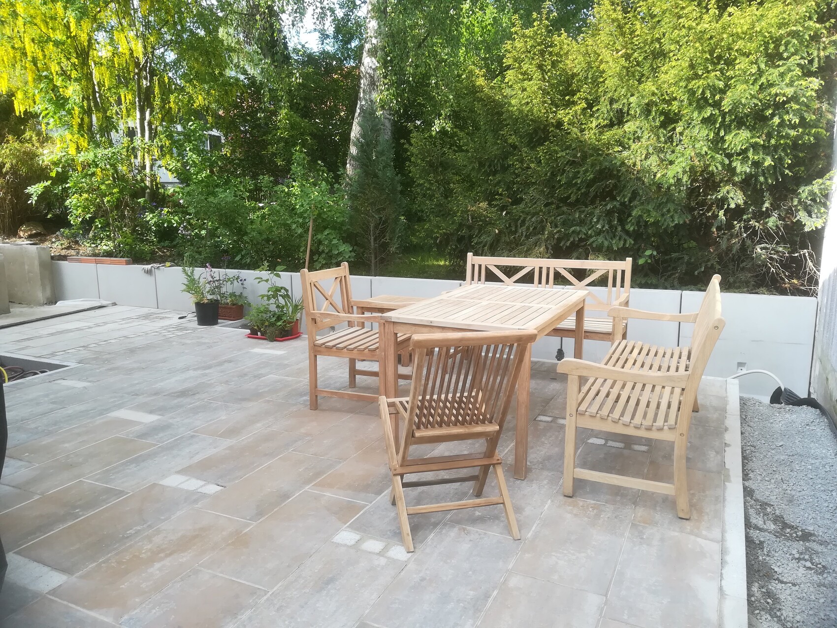 Garten-Patio mit Holztisch und Stühlen auf Terrassenplatte, grüne Hecken im Hintergrund.
