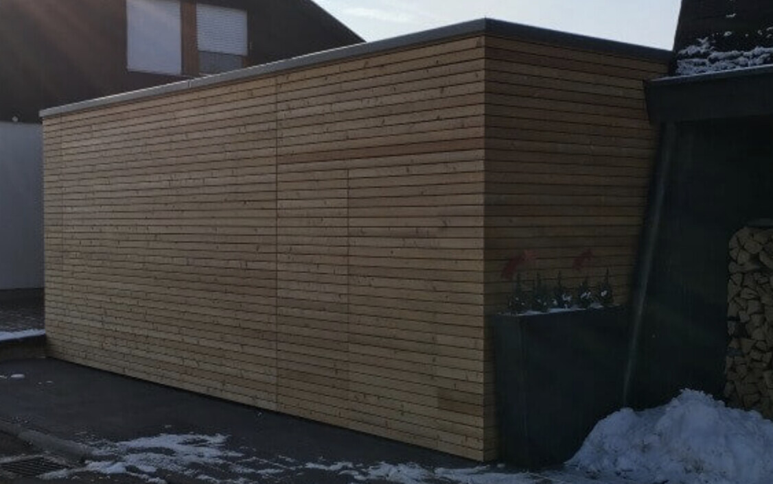 Außenwand aus horizontalen Holzlatten; rechts Pflanzkübel mit roten Blumen, Schnee.