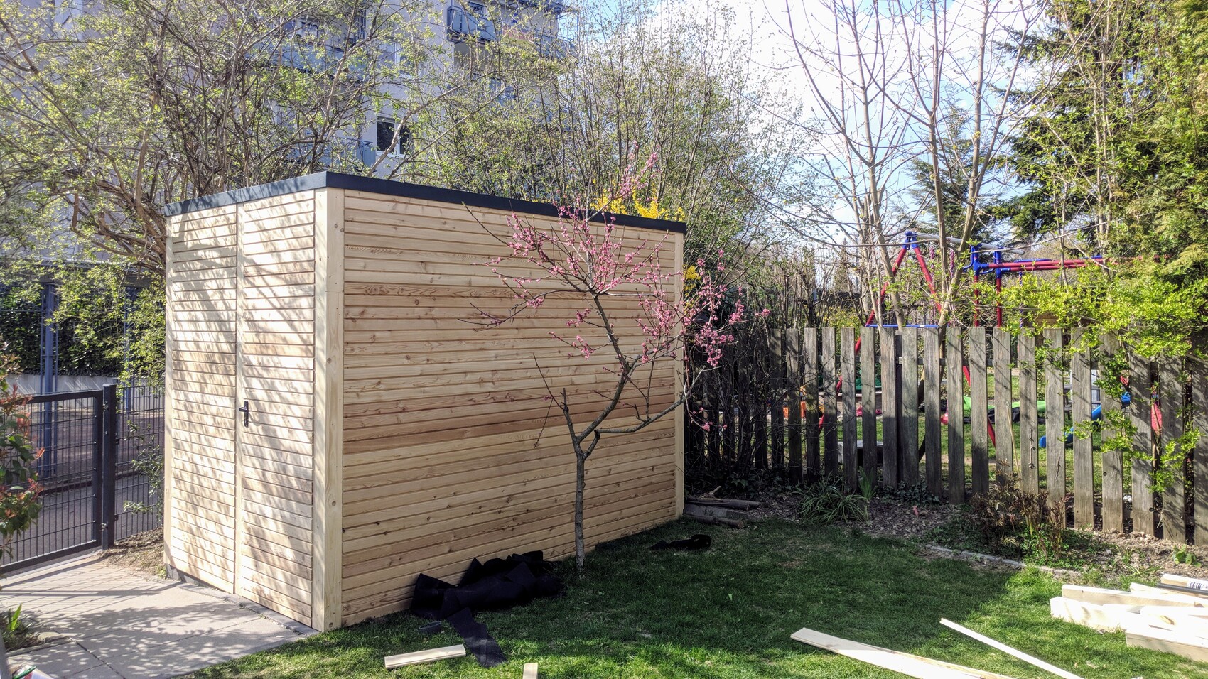 Garten mit hellem Schuppen links, blühender Baum mit rosa Blüten, Zaun rechts, Spielplatz dahinter