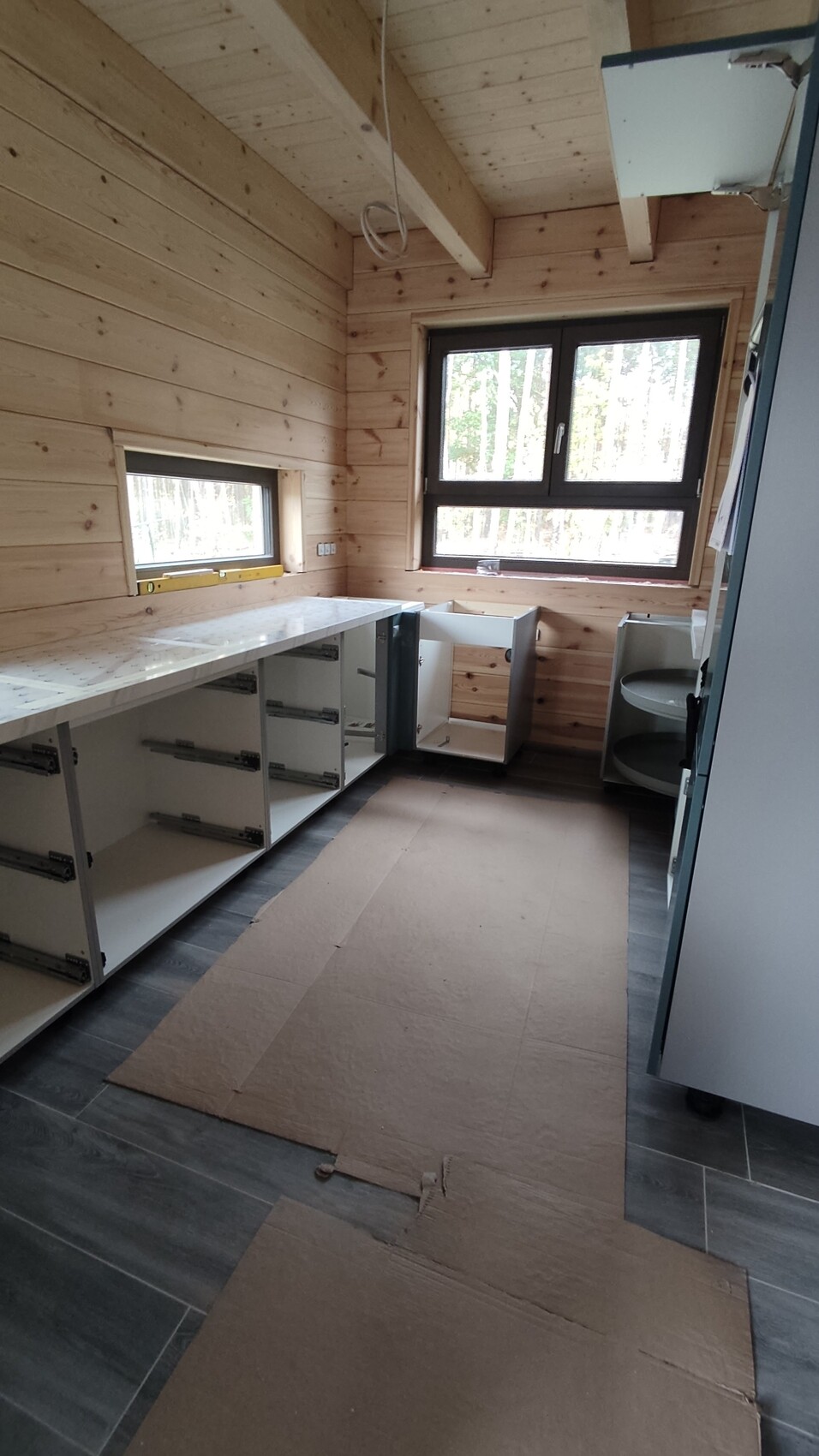 Küche in Holzbau, offene Unterschränke, Kartonboden, Fenster mit Blick auf Wald.