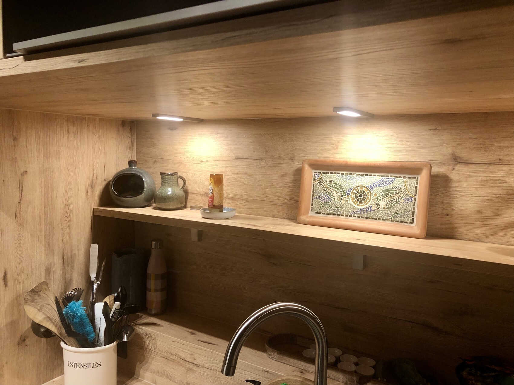 Holzküchenregal mit Keramikgefäßen, Vorratsflasche, Mosaikrahmen und Utensilien über der Spüle.