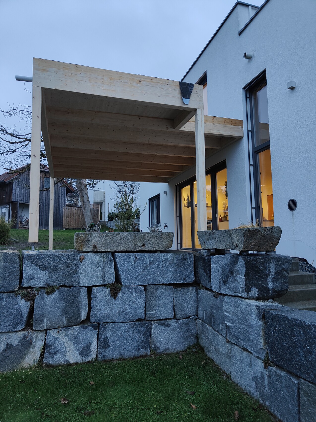Moderner Hausanbau mit Holz-Terrassendach über Steinquader-Wand und beleuchtete Innenräume.